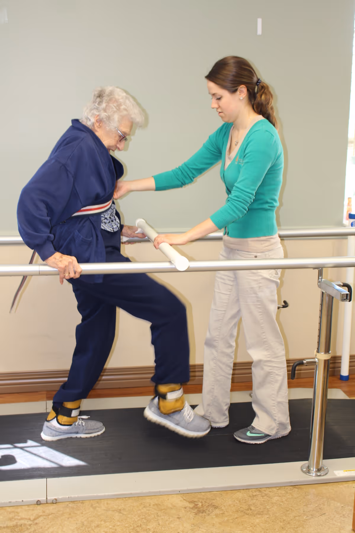An elderly woman wearing ankle weights is walking on a treadmill with parallel bars for support while a young female caregiver assists her by holding her arm and guiding her movement in a rehabilitation or physical therapy setting.