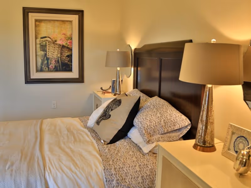 A cozy bedroom with a neatly made bed featuring patterned pillows and a decorative pillow with a bird design. The bed has a dark wooden headboard. On either side of the bed are nightstands with matching lamps. A framed picture of a bicycle with a basket of flowers hangs on the wall above one nightstand.