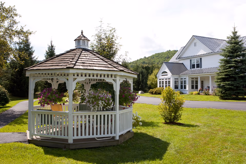 A white wooden gazebo with a shingled roof surrounded by hanging flower baskets sits on a green lawn. In the background, there is a large white building with multiple windows and a metal roof, set against a backdrop of trees and hills under a partly cloudy sky.