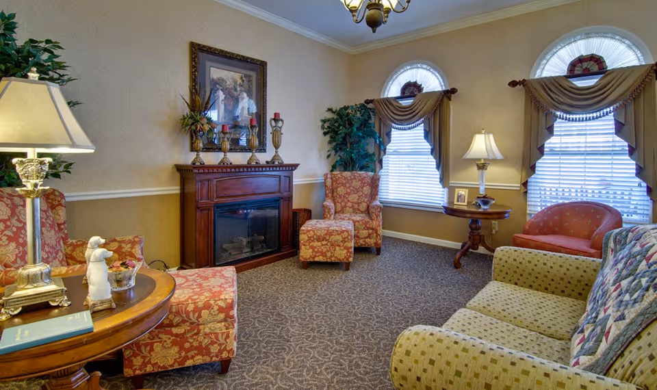 A cozy living room with patterned armchairs and a sofa arranged around a wooden electric fireplace. The room features two arched windows with decorative curtains, a side table with a lamp and framed photo, a floor lamp, and a framed painting above the fireplace. The carpet has a subtle pattern, and there are plants adding greenery to the space.
