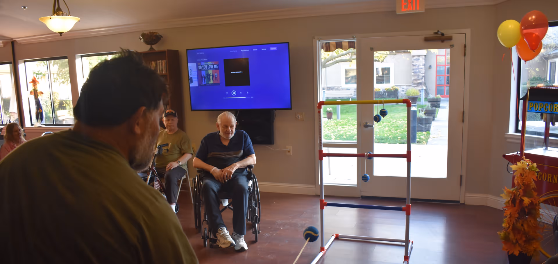 A group of elderly people in a common room at Fair Oaks Estates. One man in a wheelchair is smiling while others are seated nearby. A man in the foreground is playing a game involving tossing balls onto a ladder-like structure. The room has large windows, a TV mounted on the wall, and a popcorn machine decorated with balloons and autumn leaves.
