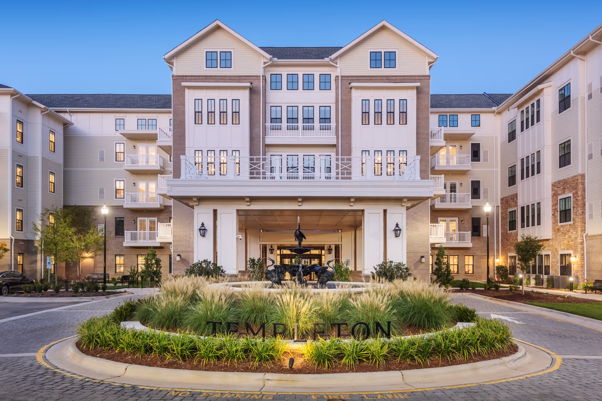 Front exterior view of The Templeton of Cary, a multi-story senior living facility with a central entrance, balconies, and a landscaped roundabout featuring a fountain and greenery.
