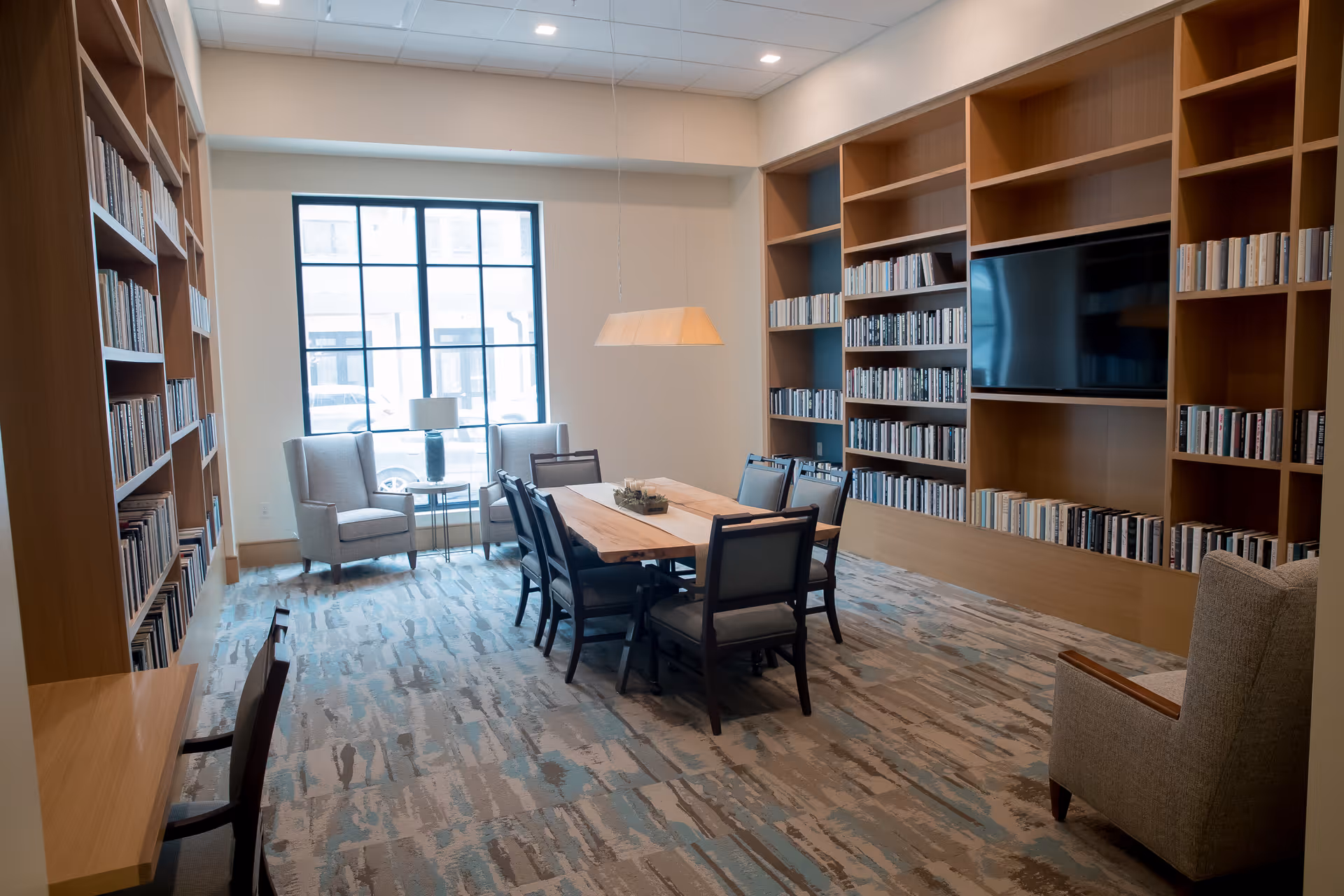 A well-lit room with large bookshelves filled with books on three walls, a wooden table with six chairs in the center, two armchairs near a large window, and a flat-screen TV mounted on one bookshelf.