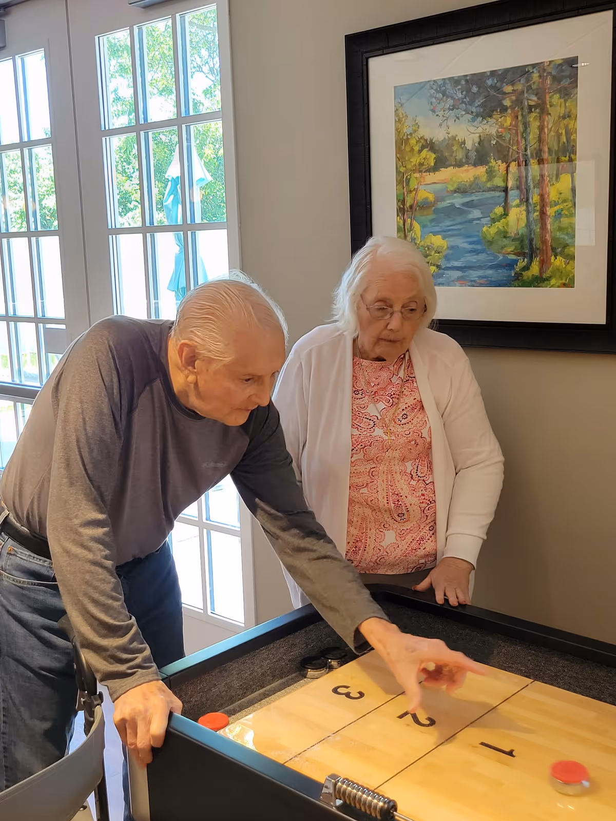 An elderly man and woman playing shuffleboard indoors near large windows with natural light. The man is leaning over the shuffleboard table, aiming to slide a puck, while the woman watches attentively. A framed painting of a river and trees hangs on the wall behind them.