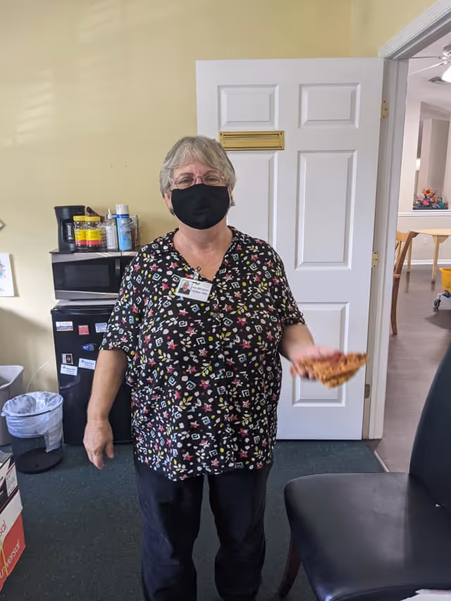 A woman wearing a black face mask and a patterned blouse stands indoors holding a slice of pizza. Behind her is a white door, a small black refrigerator with various items on top, and a trash can. Part of a chair and another room with tables and chairs are visible through an open doorway.