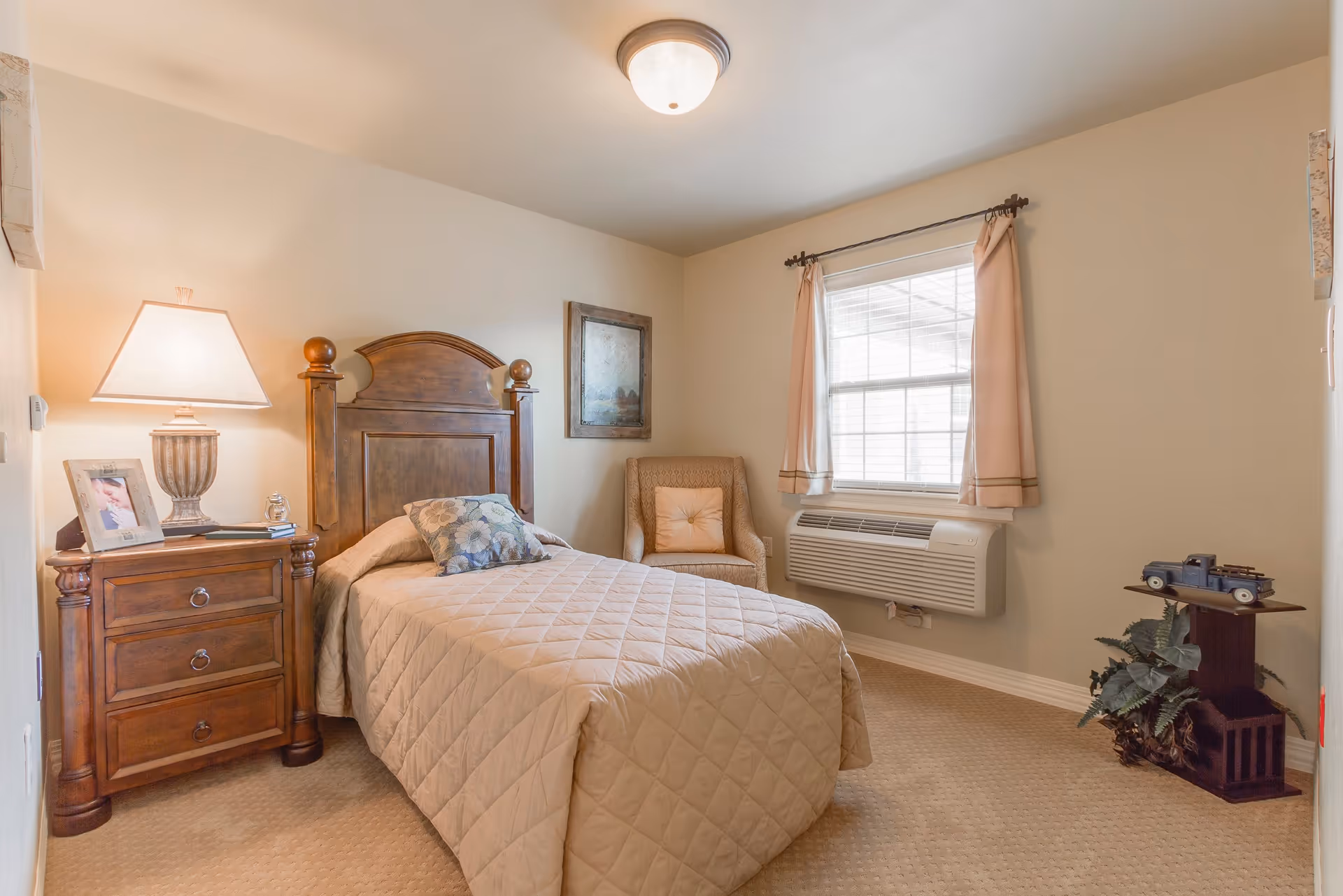 A cozy bedroom with a single bed covered in a beige quilted bedspread and a floral pillow. Next to the bed is a wooden nightstand with three drawers, a table lamp, a framed photo, and some books. There is a cushioned armchair with a pillow near a window with beige curtains. Below the window is a wall-mounted air conditioning unit. A small wooden stand with a decorative model truck and a plant is in the corner. The walls are light-colored, and the carpet is beige.