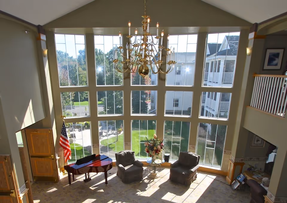Bright and spacious living room area with large floor-to-ceiling windows letting in natural light, two armchairs around a small round table with a flower arrangement, a grand piano near an American flag, and a large chandelier hanging from the ceiling.