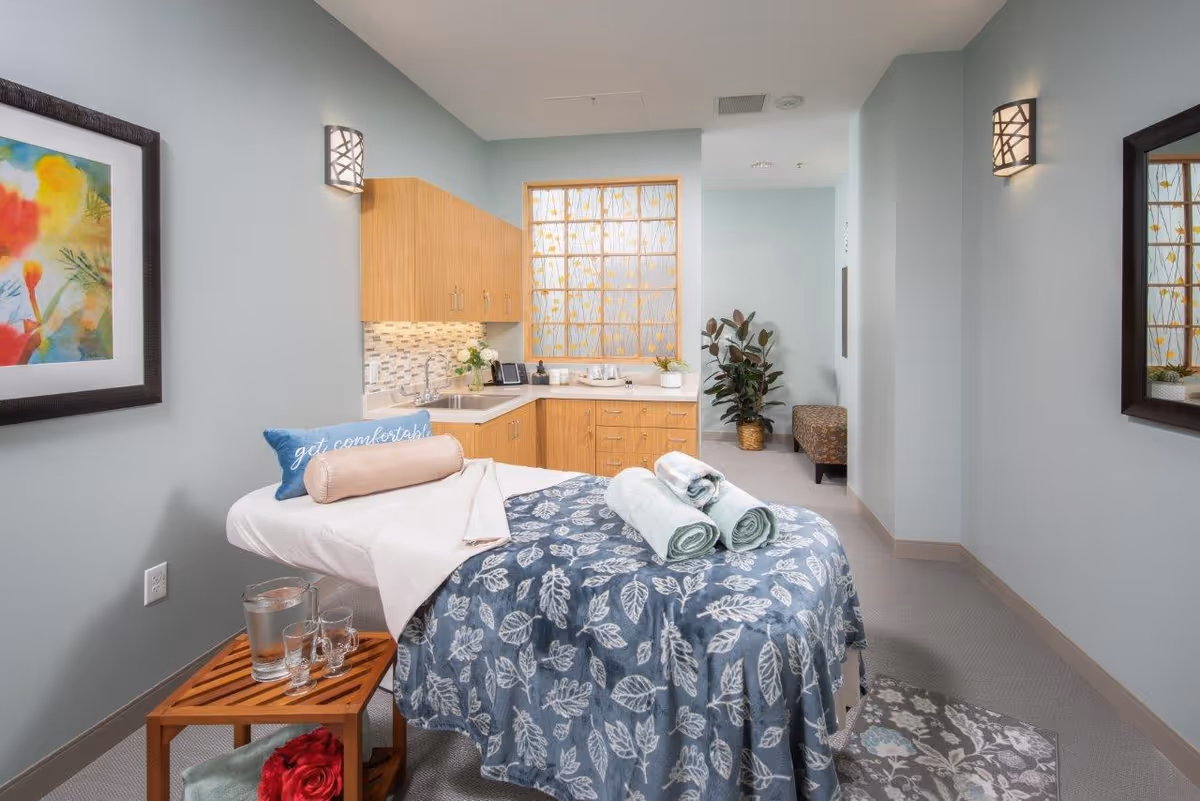 A cozy massage or therapy room with a massage table covered in a blue leaf-patterned blanket and rolled towels on top. There is a small wooden side table with a pitcher of water and glasses. The room has light blue walls, wooden cabinets with a sink, a colorful framed painting on one wall, a large decorative window, and a potted plant in the corner. Soft lighting fixtures are mounted on the walls.