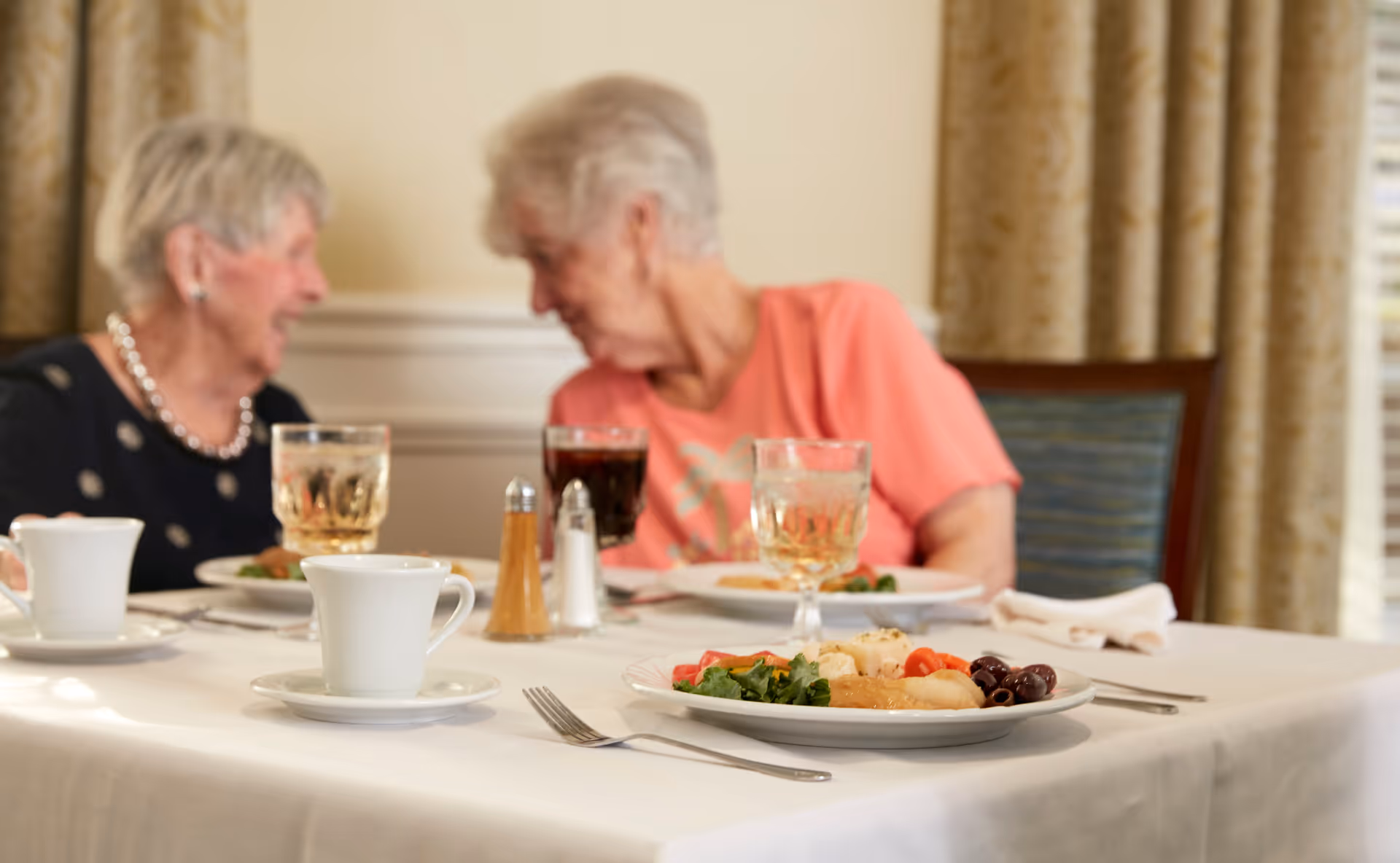 Two elderly women sitting at a dining table with plates of food, glasses of beverages, and coffee cups, engaged in conversation in a warmly lit room with curtains in the background.
