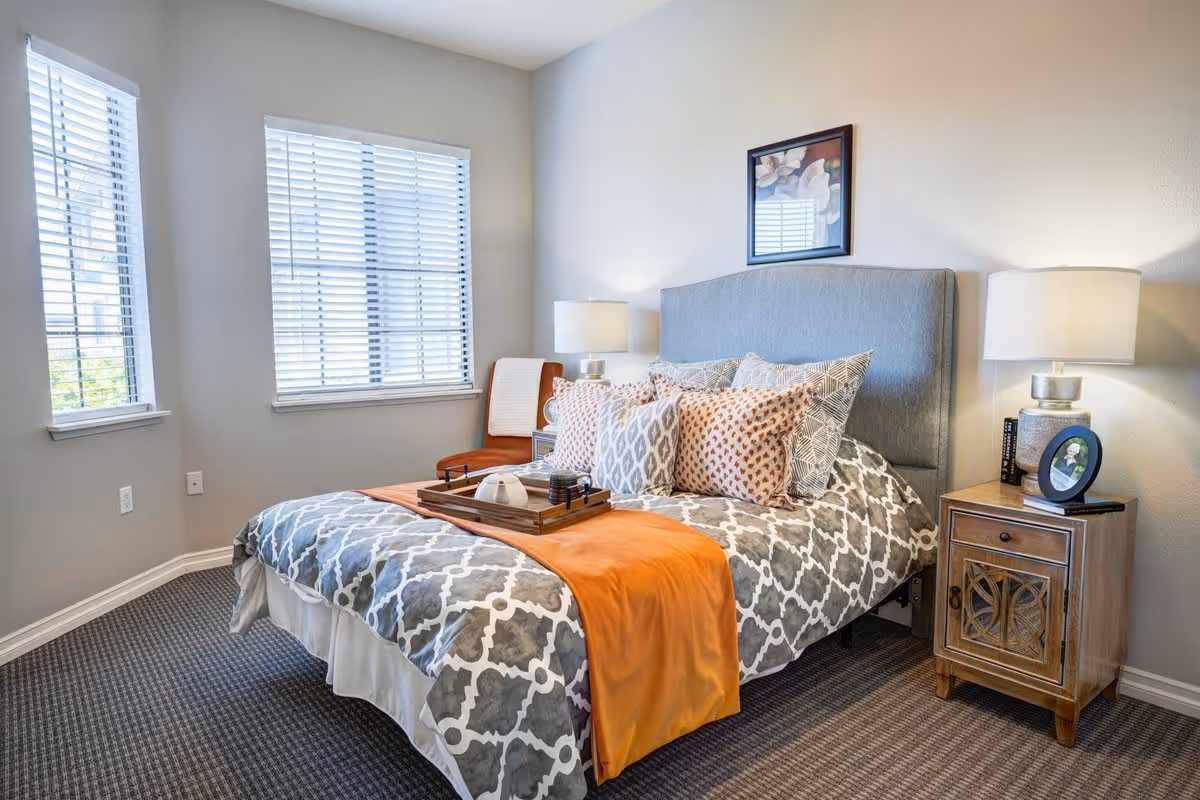 A well-lit bedroom with a large bed featuring a gray and white patterned comforter, multiple decorative pillows, and an orange throw blanket. There is a wooden nightstand with a lamp, books, and a framed photo on the right side of the bed. Two windows with white blinds allow natural light into the room. A framed floral picture hangs above the bed, and a chair with a blanket is positioned near the windows.