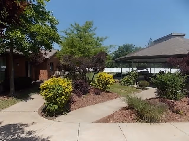 Outdoor garden area with a paved walkway, green bushes, trees, and a covered pavilion with seating. A brick building is partially visible on the left side under a clear blue sky.