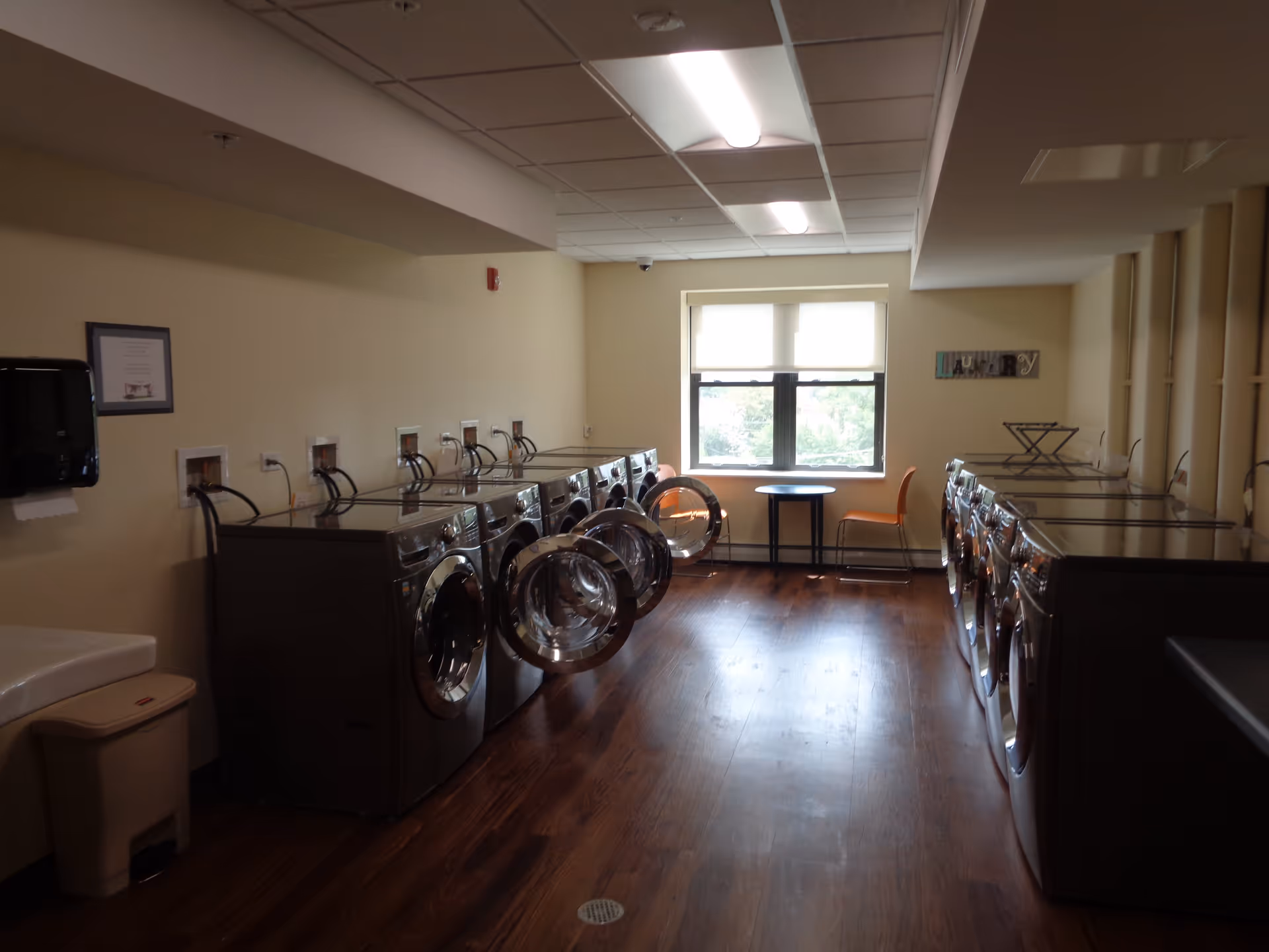 Laundry room with two rows of front-loading washing machines and dryers, some with doors open. There is a window at the far end with a small round table and two orange chairs. The room has wood flooring and beige walls with a sign that says 'Laundry'.