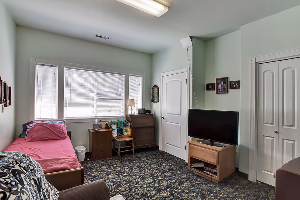 A small residential room with a single bed covered in a pink sheet and pillow, a wooden nightstand with a lamp and other items, a wooden chair with a colorful crocheted blanket, a wooden dresser, a flat-screen TV on a wooden stand, and framed photos on the light green walls. The room has a patterned carpet and a large window with blinds letting in natural light.