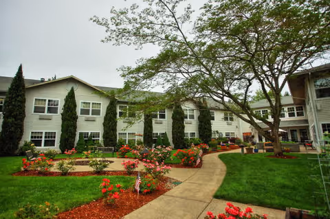 A landscaped garden area with a paved walkway winding through green grass and flower beds filled with red and pink flowers. Tall trees and shrubs are present, and a two-story building with many windows surrounds the garden area under an overcast sky.