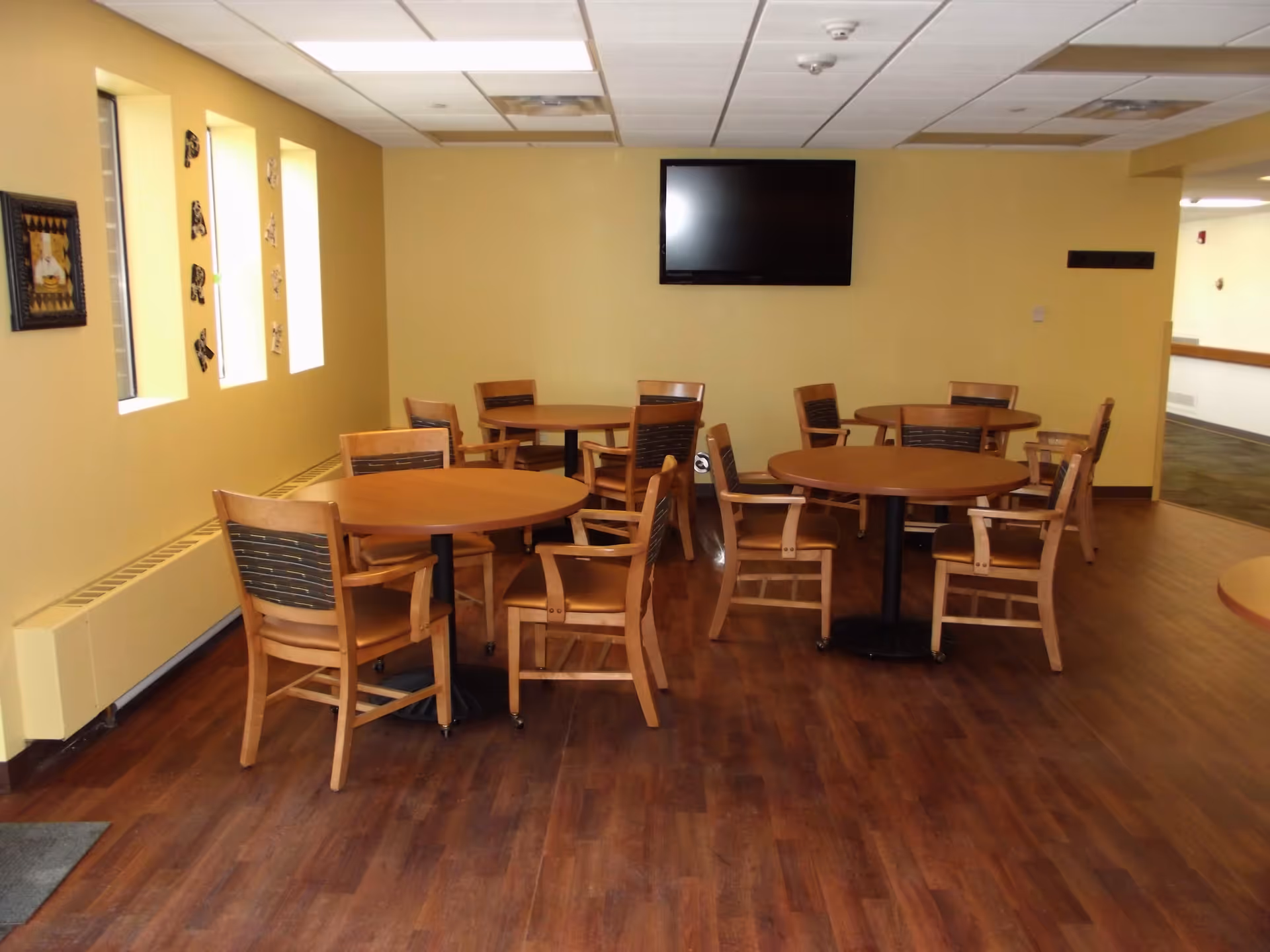 Communal dining room with round wooden tables and chairs, a wall-mounted TV, and yellow walls.
