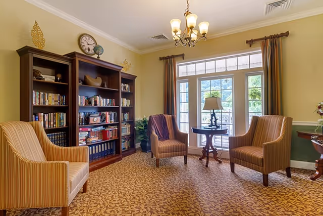 A cozy living room area with three striped armchairs arranged around a small round wooden table with a lamp. Behind the chairs is a large bookshelf filled with books and decorative items. The room has a patterned carpet, yellow walls, a chandelier, and large glass doors with curtains leading outside.
