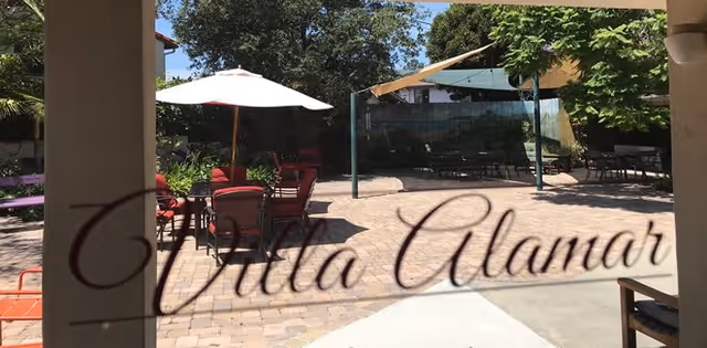 Outdoor patio with tables, chairs, umbrellas and a shaded pergola seen through glass with the words 'Villa Alamar' scripted across it.