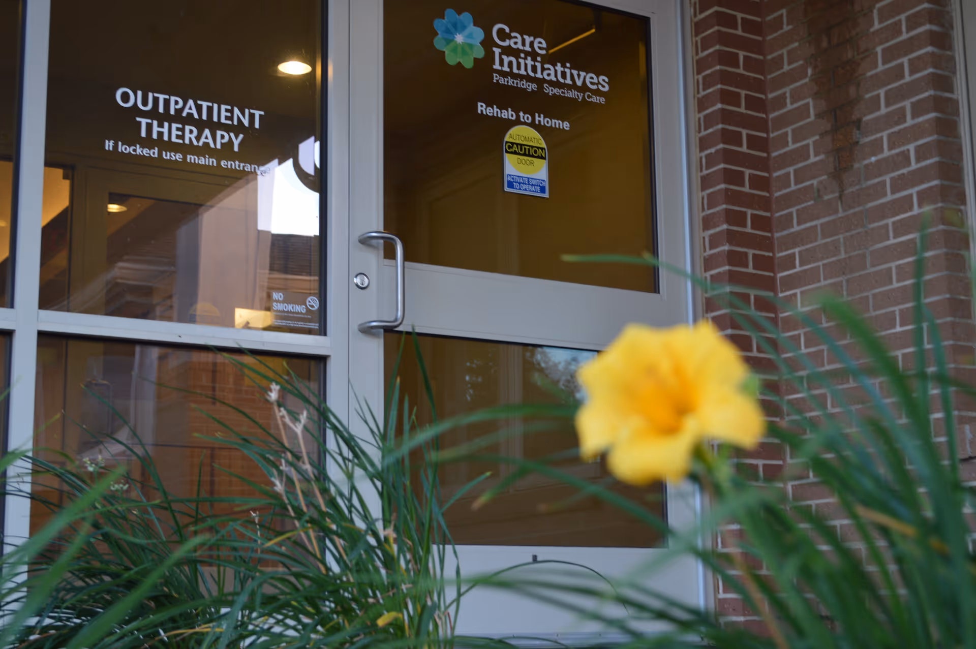 Entrance door to the outpatient therapy area at Parkridge Nursing & Rehab Center with signage for Care Initiatives Parkridge Specialty Care and a yellow flower and green plants in the foreground.