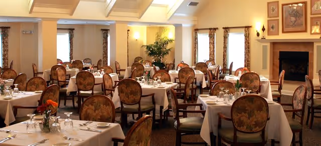 A spacious dining room with multiple tables covered in white tablecloths, each set with glassware, silverware, and floral centerpieces. The room features upholstered chairs with floral patterns, large windows with curtains, warm lighting, and a fireplace on the right wall.