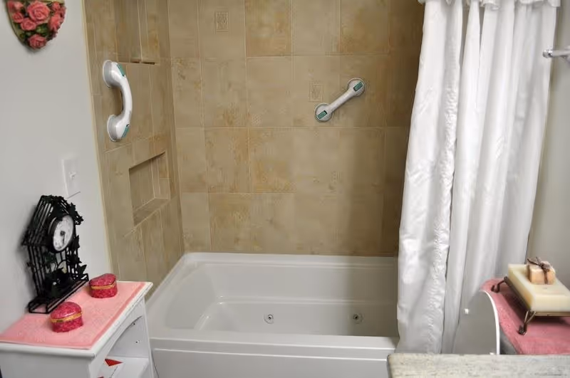 A bathroom featuring a white bathtub with beige tiled walls and two white grab bars. A white shower curtain is partially drawn to the right. On the left, there is a small white shelf with a black decorative clock and two small pink containers on a pink mat. On the right, a countertop with a soap dish holding soap is visible.