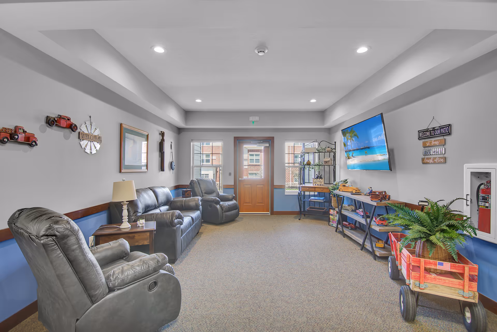 A cozy senior living common area with three black leather recliners, a wooden side table with a lamp, wall decorations including red toy trucks and a welcome sign, a large flat-screen TV mounted on the wall displaying a beach scene, a shelving unit with various items, a red wagon with a potted plant, and a door with windows letting in natural light.