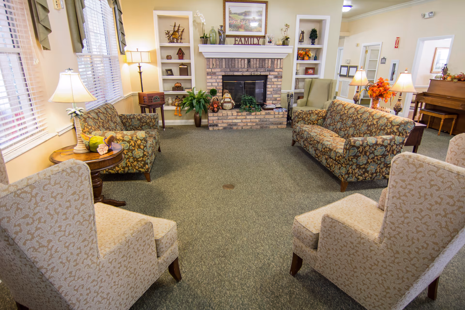 A cozy living room area with two patterned sofas, two beige armchairs, a brick fireplace with decorative items and plants on the mantel and shelves, a wooden piano in the background, and several lamps providing warm lighting.