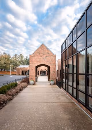 Paved walkway leading to a brick arched entrance with large glass windows along the right and planters under a cloudy sky.
