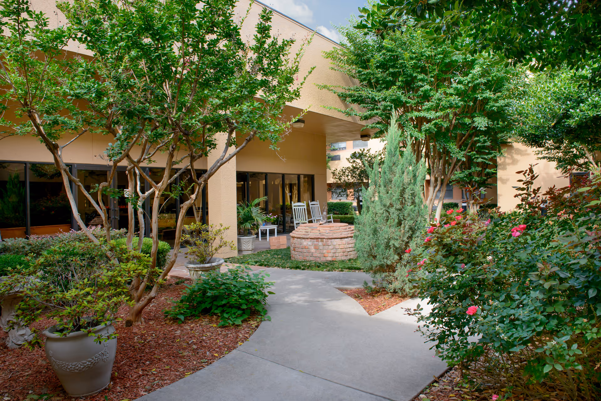 A peaceful outdoor courtyard at Renaissance Park Multi Care Center featuring a paved walkway surrounded by lush greenery, flowering bushes, and potted plants. There are two white rocking chairs near a circular brick structure under a covered area attached to the building.