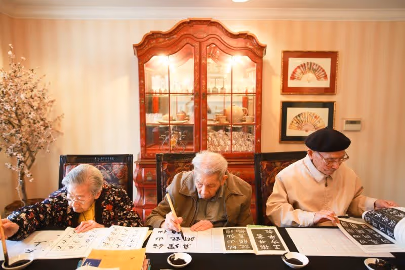 Three elderly individuals seated at a table engaged in calligraphy, with brushes and ink on paper. Behind them is a wooden cabinet with glass doors displaying glassware and dishes, and framed artwork on the wall.