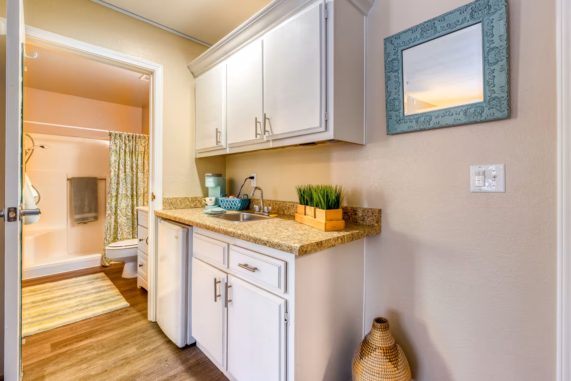 Interior view of a small kitchenette area with white cabinets, a countertop with a sink, a mini refrigerator, a basket with cups, and a decorative plant. To the left, there is an open door leading to a bathroom with a shower curtain, toilet, and a striped rug on the floor. A decorative mirror with a blue frame hangs on the wall to the right.