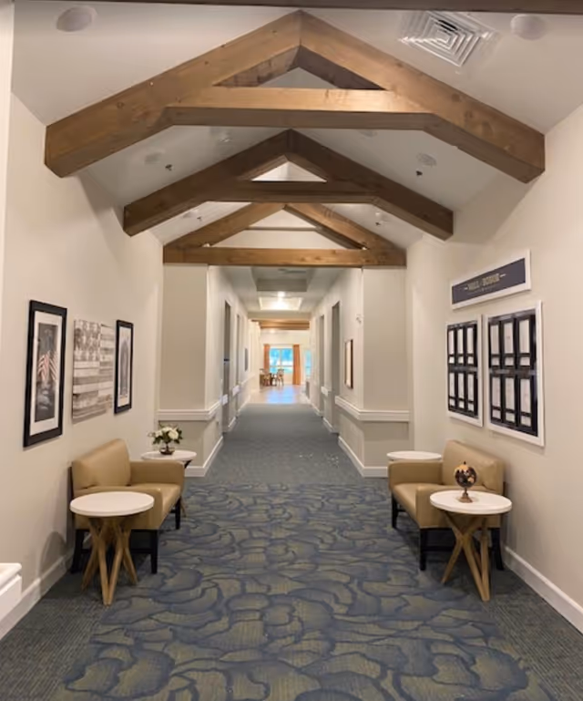 A long, carpeted hallway with wooden beams on the ceiling. There are beige chairs and small round tables with decorative items on both sides of the hallway. The walls are adorned with framed pictures and a bulletin board. The hallway leads to a bright area with windows at the far end.