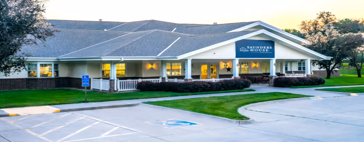 Exterior view of Saunders House, a single-story building with a covered porch, white railings, and a sign displaying the facility name. The building is surrounded by green grass and trees, with a parking lot and handicap parking spaces in the foreground.