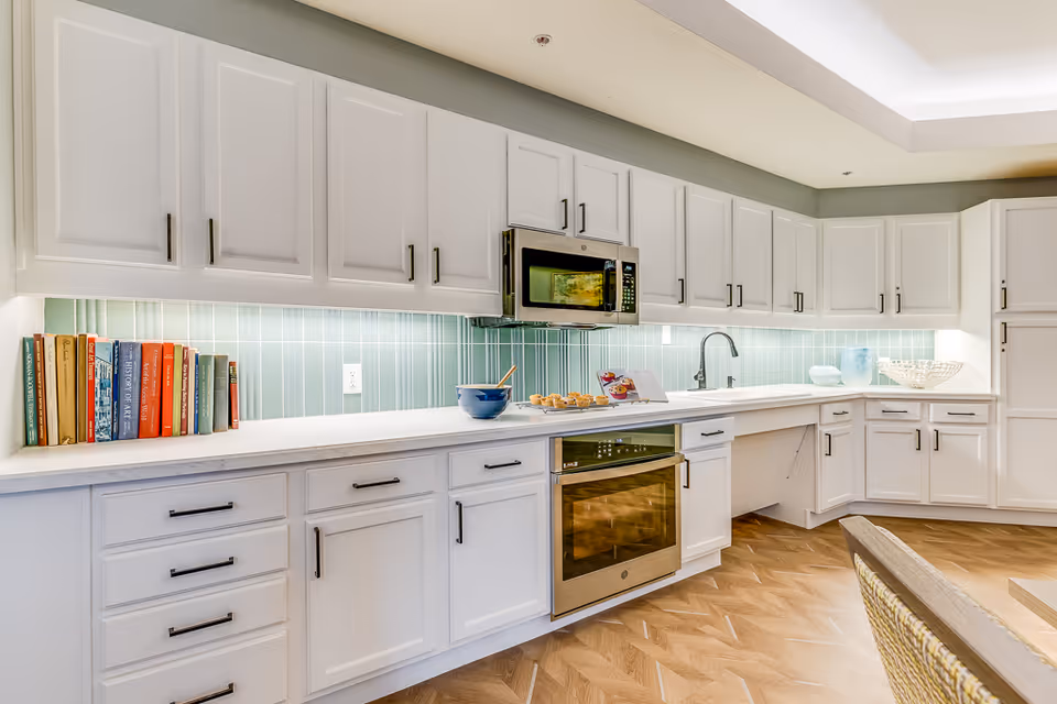 Bright and modern kitchen with white cabinets, a light blue tiled backsplash, stainless steel microwave and oven, a sink, and a countertop with cookbooks, a bowl, muffins, and a cookbook stand.