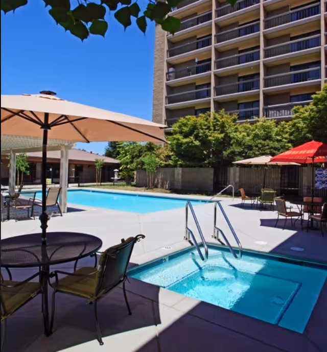 Outdoor pool area with a hot tub in the foreground, surrounded by patio tables with umbrellas and chairs. A multi-story building and trees are visible in the background under a clear blue sky.