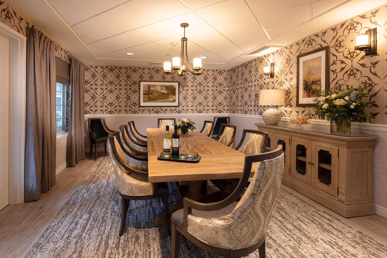 A warmly lit dining room with a long wooden table surrounded by upholstered chairs. The walls feature patterned wallpaper and framed artwork. A sideboard with a lamp, flowers, and a plate of cookies is against one wall. A chandelier hangs above the table, which has two bottles of wine and glasses on a tray.