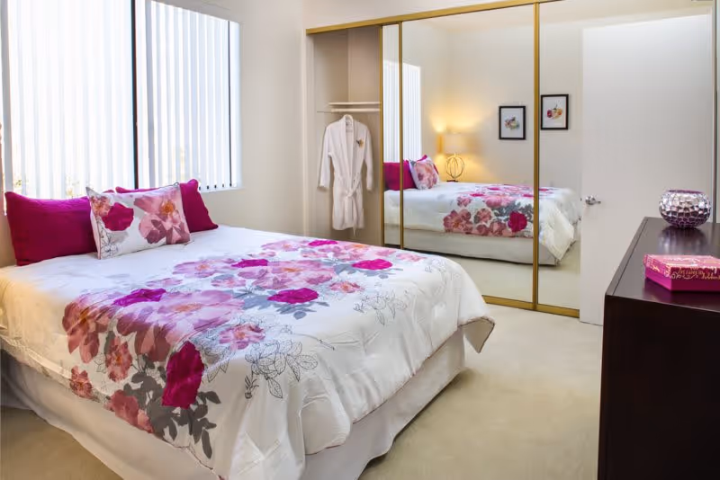A bright bedroom with a bed covered in a white comforter featuring pink and purple floral patterns, two magenta pillows, and a floral accent pillow. There is a large window with vertical blinds on the left wall, a mirrored closet with gold trim on the right, and a dark wooden dresser with a decorative purple candle holder and a pink box on top. A white robe hangs inside the closet, and two framed floral pictures are on the wall next to a lit lamp.