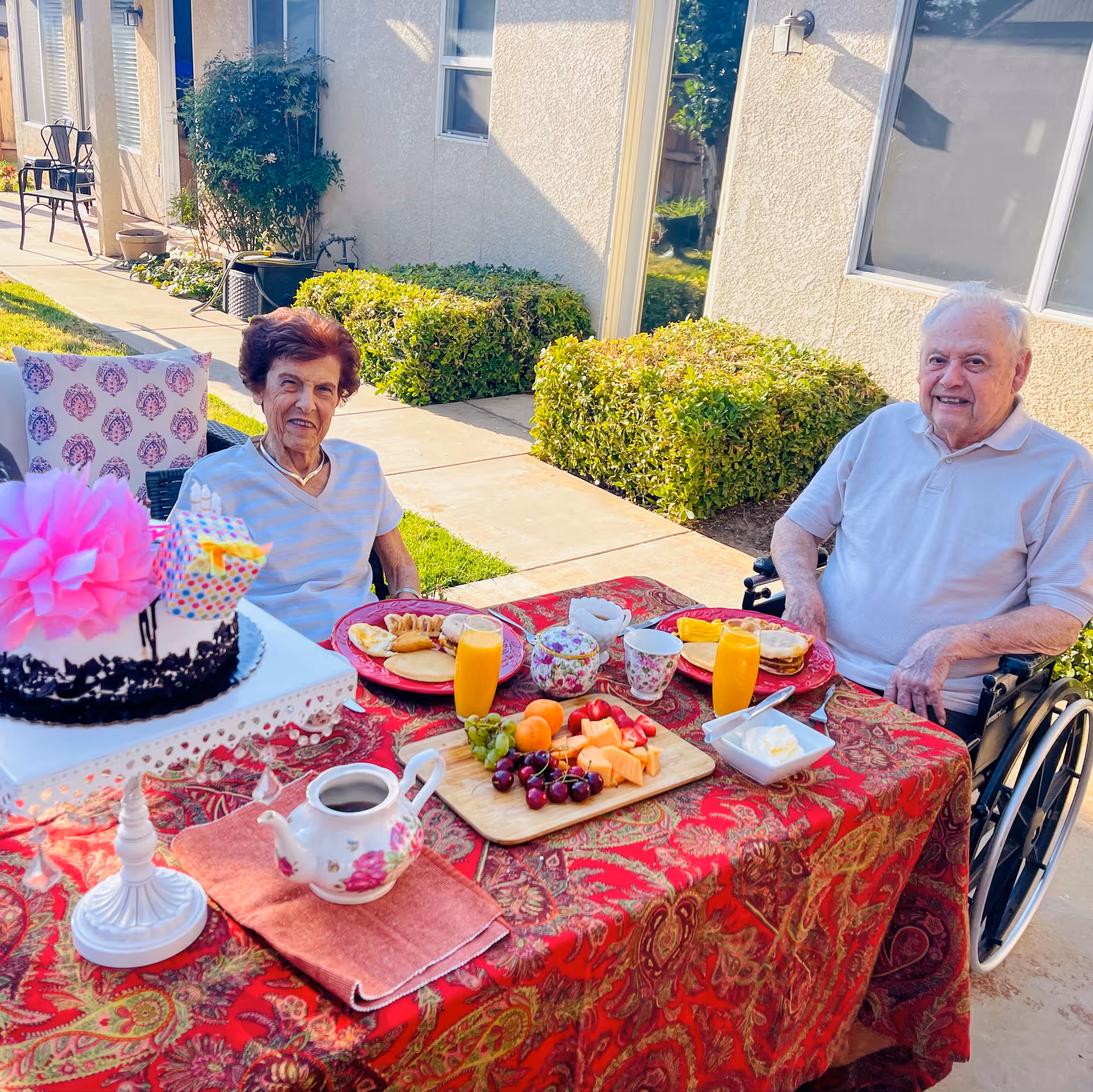 Two elderly individuals, one woman and one man in a wheelchair, sitting at a table outdoors with a red patterned tablecloth. The table is set with breakfast items including plates with food, glasses of orange juice, a teapot, a cutting board with assorted fruits, and a decorated cake with a large pink flower on top. The setting is a sunny patio area with bushes and a building wall in the background.
