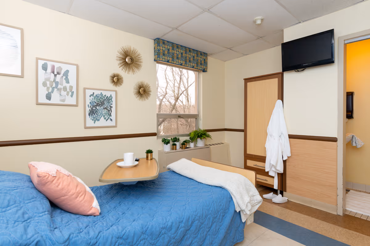 A cozy nursing center bedroom with a blue quilted bedspread and a pink pillow. A small overbed table holds a white cup and saucer. The room has a window with a patterned valance and several small potted plants on the windowsill. Three decorative wall hangings and two framed abstract art pieces adorn the wall. A white robe and slippers hang near a wooden cabinet, and a flat-screen TV is mounted on the wall above the cabinet. The door to a bathroom is partially visible.