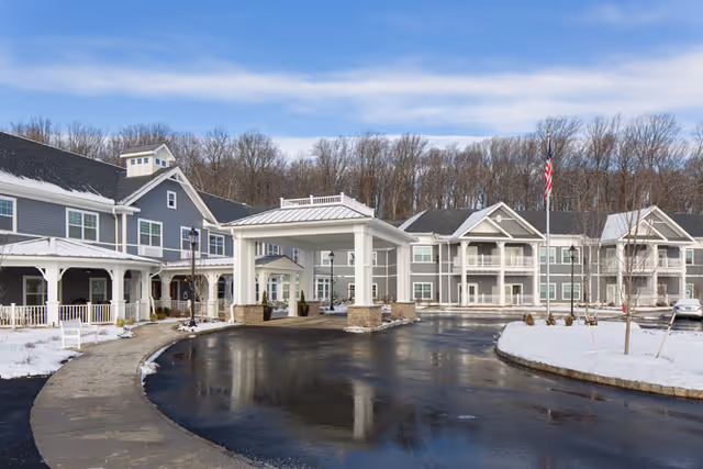 Exterior view of Brightview Warren - Senior Assisted Living & Memory Care facility on a clear winter day with snow on the ground, a circular driveway, and an American flag in front of the building surrounded by trees.