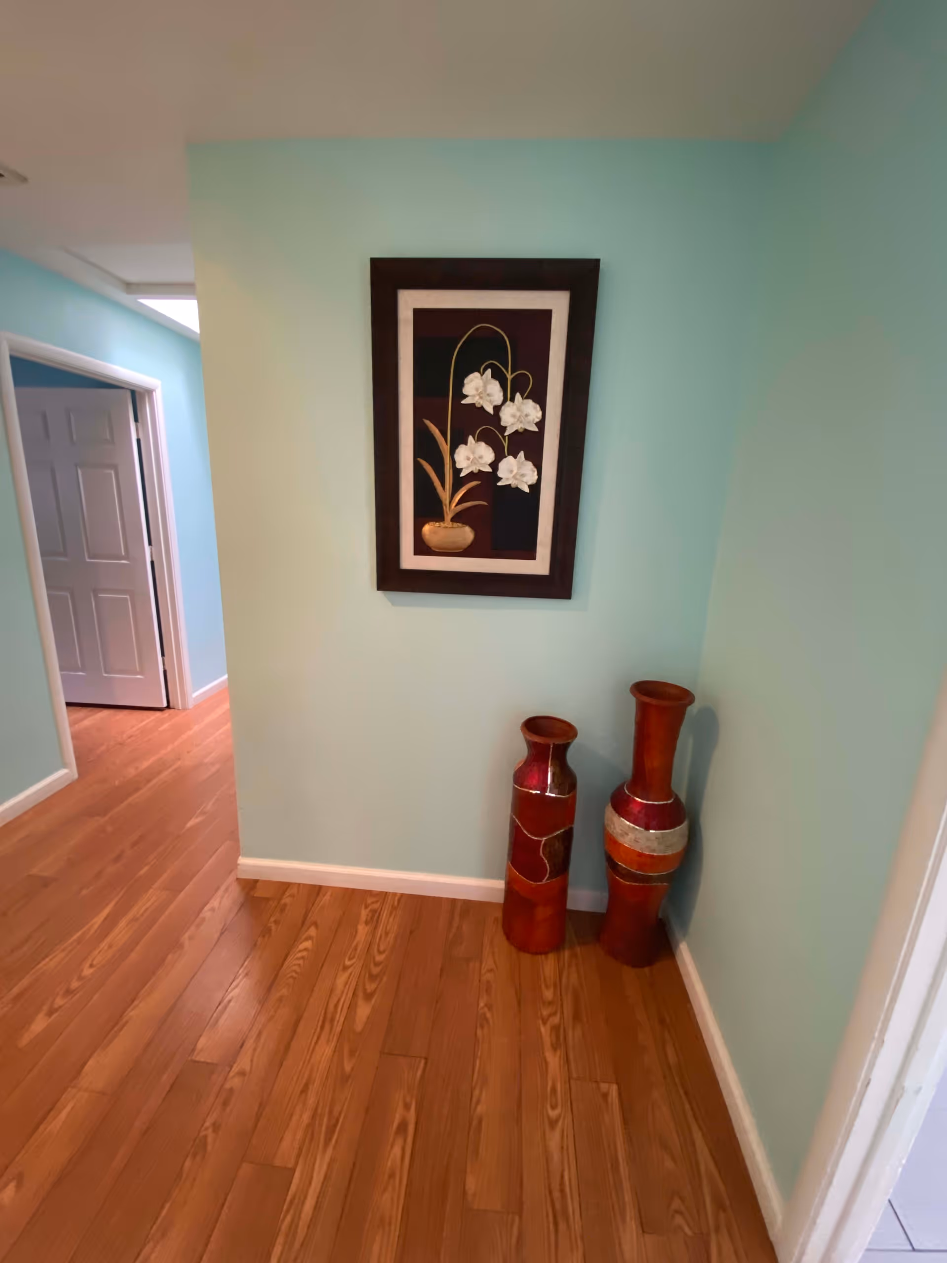 A hallway with light blue walls and wooden flooring. On the right wall, there is a framed artwork featuring white flowers in a brown pot. Below the artwork, two tall decorative vases with red and brown tones are placed on the floor. An open doorway is visible on the left side of the hallway.