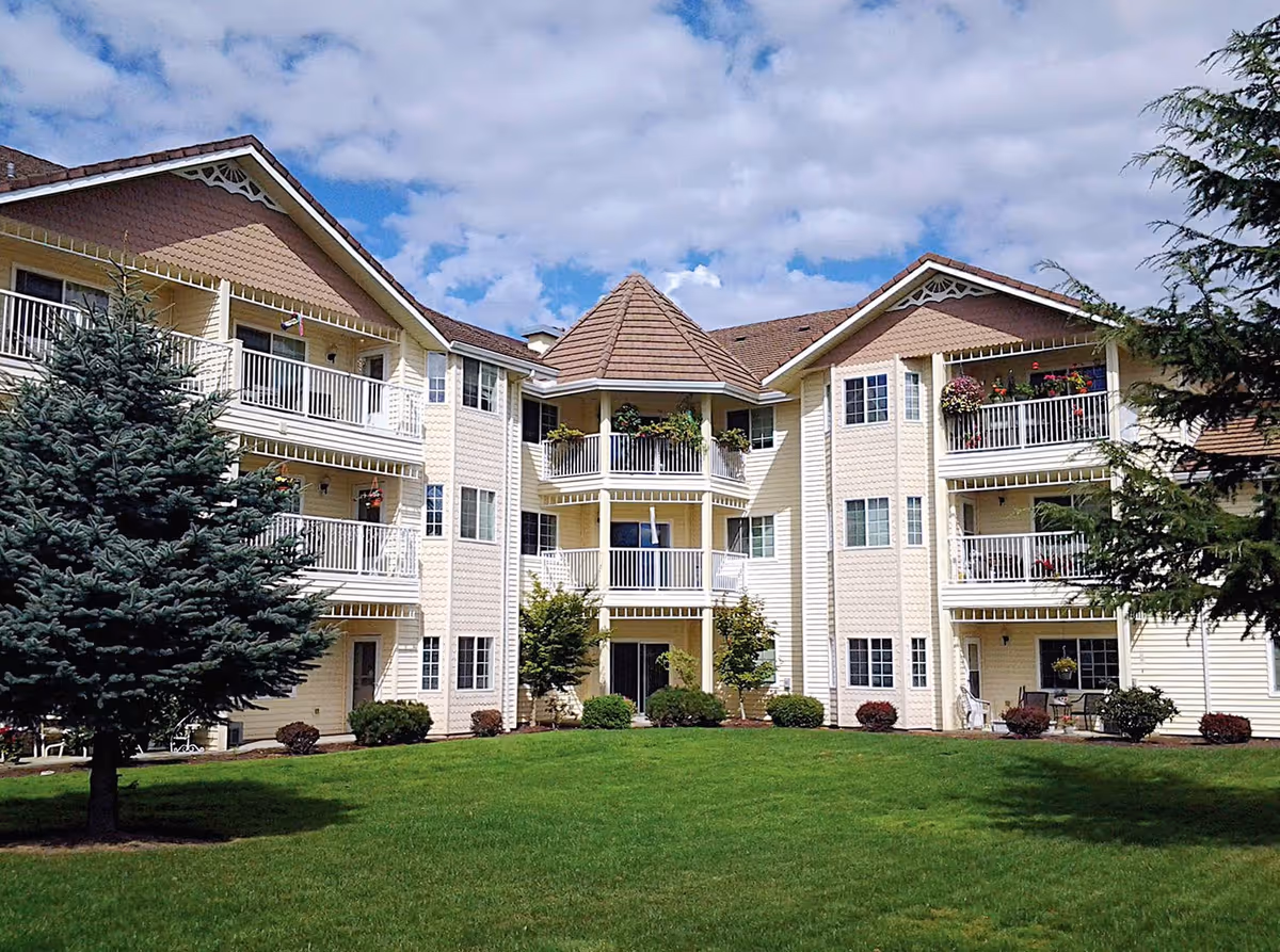 Exterior view of a multi-story retirement community building with balconies decorated with plants and flowers. The building is surrounded by a well-maintained green lawn and trees under a partly cloudy sky.