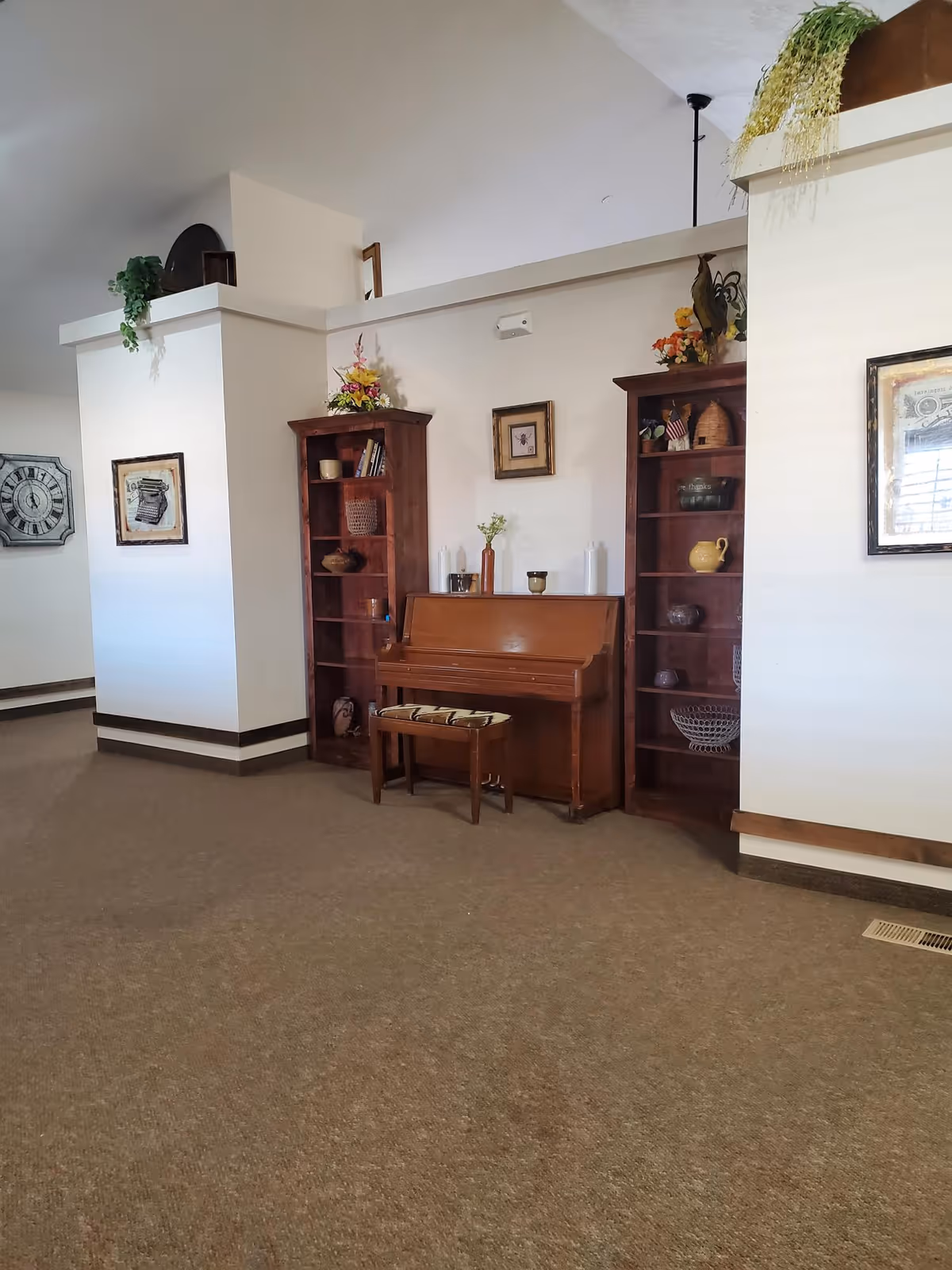 Interior view of a room with a wooden piano and matching bench placed between two wooden bookshelves filled with decorative items and books. The walls are light-colored with framed pictures hanging, and there are plants and flowers on top of the shelves. The floor is carpeted, and the ceiling is vaulted.