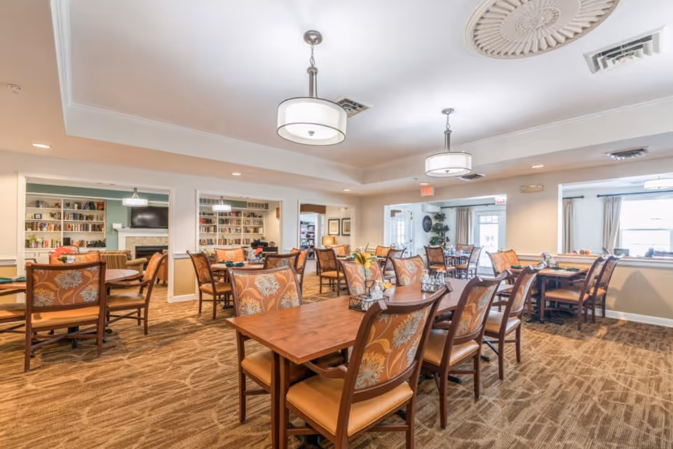 Bright communal dining room with multiple wooden tables and upholstered chairs, pendant lights, and bookshelves in the background.