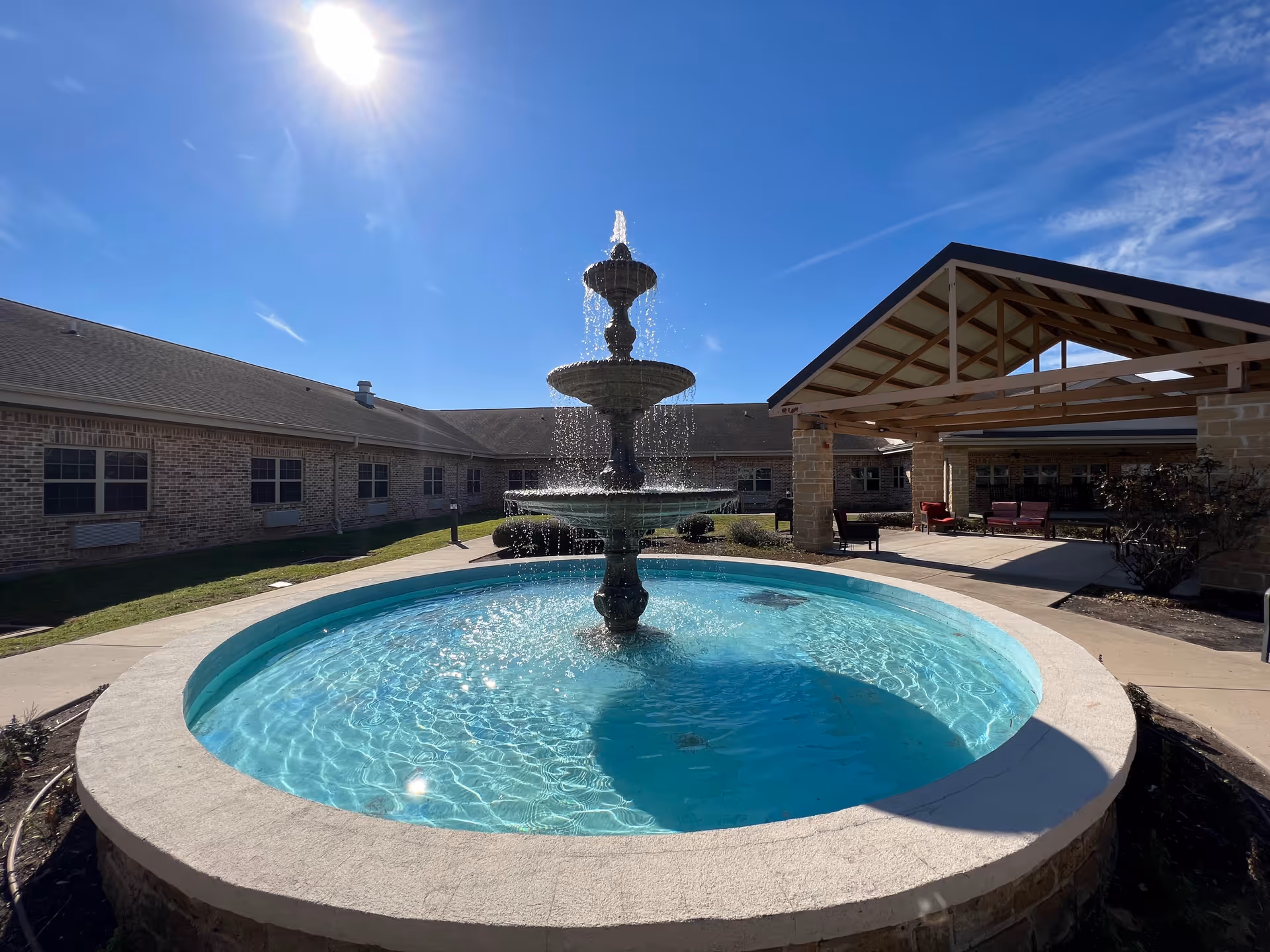 Outdoor courtyard area at Capstone Healthcare Estates on Orem featuring a large, multi-tiered water fountain in the center of a circular pool. Surrounding the fountain are paved walkways, a covered seating area with chairs and benches, and a brick building under a clear blue sky with the sun shining brightly.
