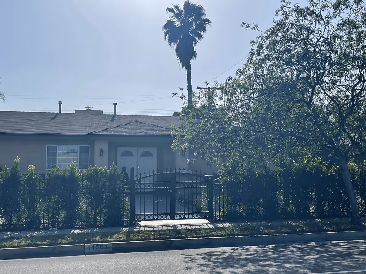 Front exterior view of a single-story residential building with a gated fence, bushes, a palm tree, and other trees in front. The building has a double door entrance and a window with bars on the left side.