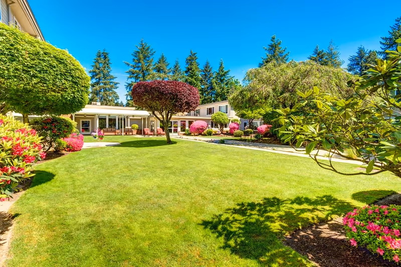 Sunny landscaped courtyard with a green lawn, trimmed ornamental trees and flowering shrubs bordered by the senior living building.