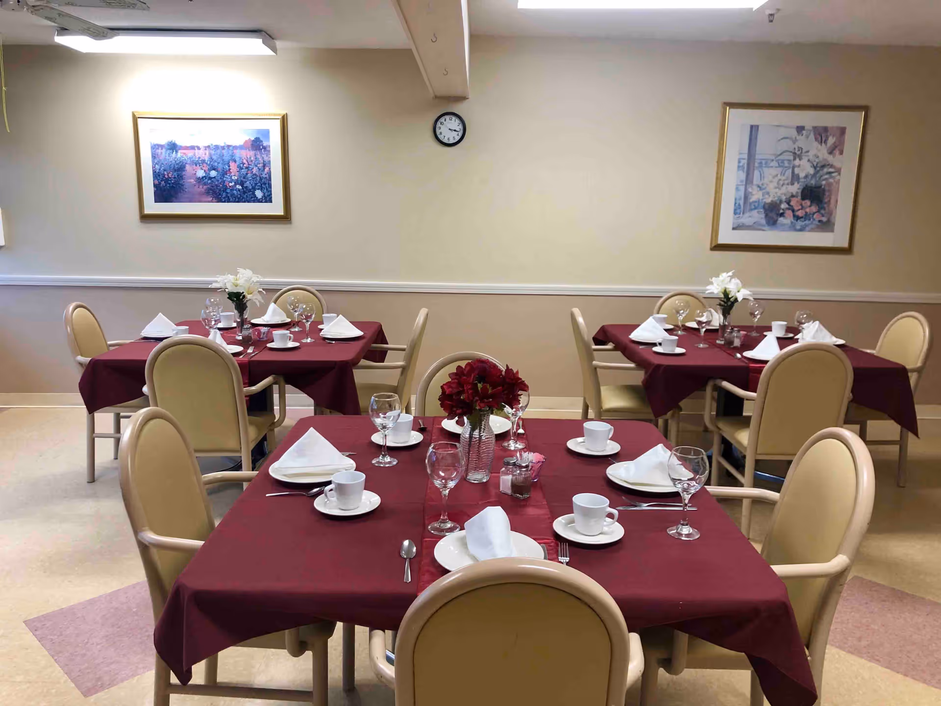 Dining room with four tables covered in burgundy tablecloths, each set with white napkins, plates, cups, wine glasses, and floral centerpieces. Beige chairs surround the tables. Two framed floral paintings hang on the beige walls, and a clock is visible above one of the paintings.