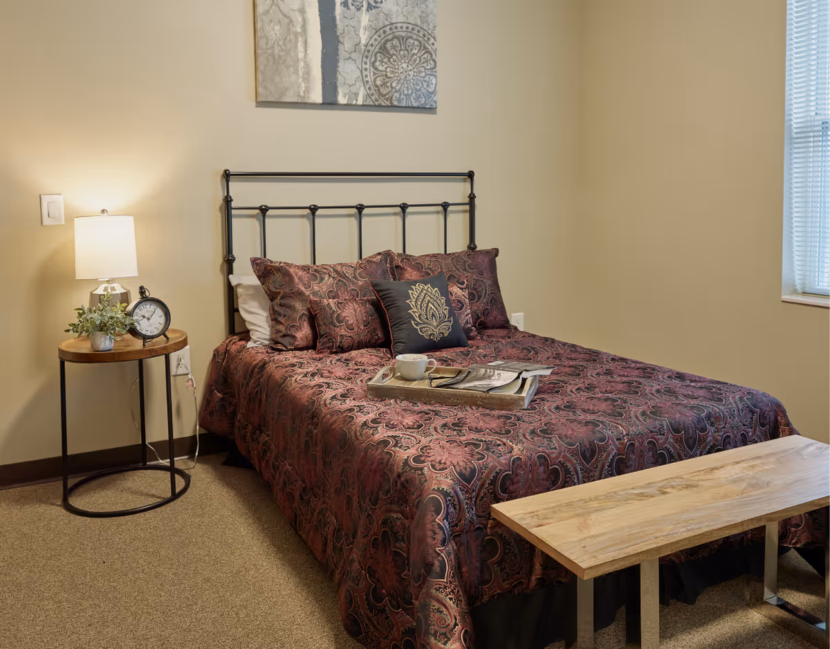 A tidy bedroom with a patterned bedspread and metal headboard, a side table with a lamp and clock, and a wooden bench at the foot of the bed.
