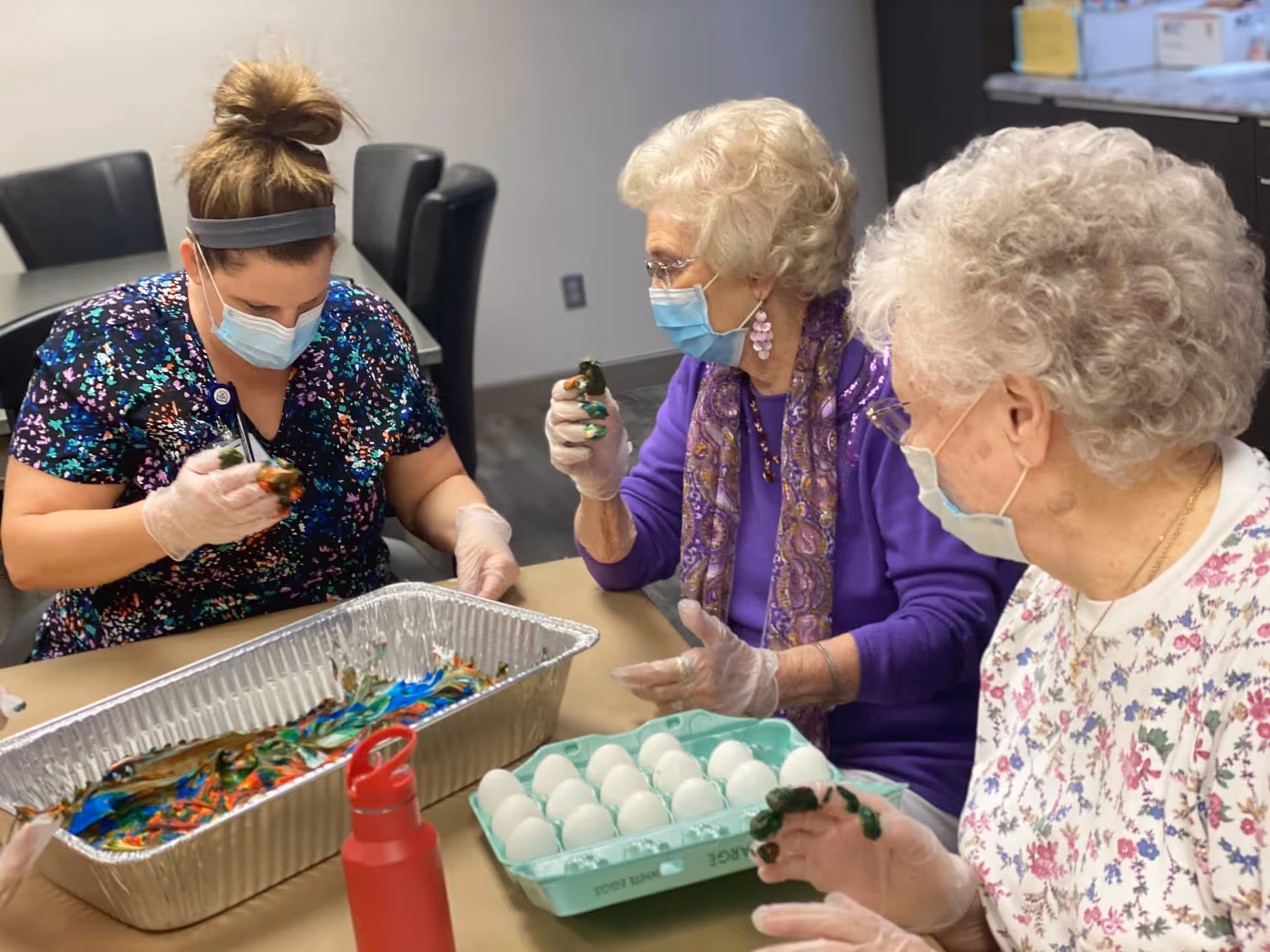 Three women wearing face masks and gloves are sitting around a table engaged in an egg decorating activity. One woman is holding a painted egg over a tray filled with colorful paint, while another woman holds an egg carton with white eggs. They appear to be in a communal room with chairs and tables in the background.