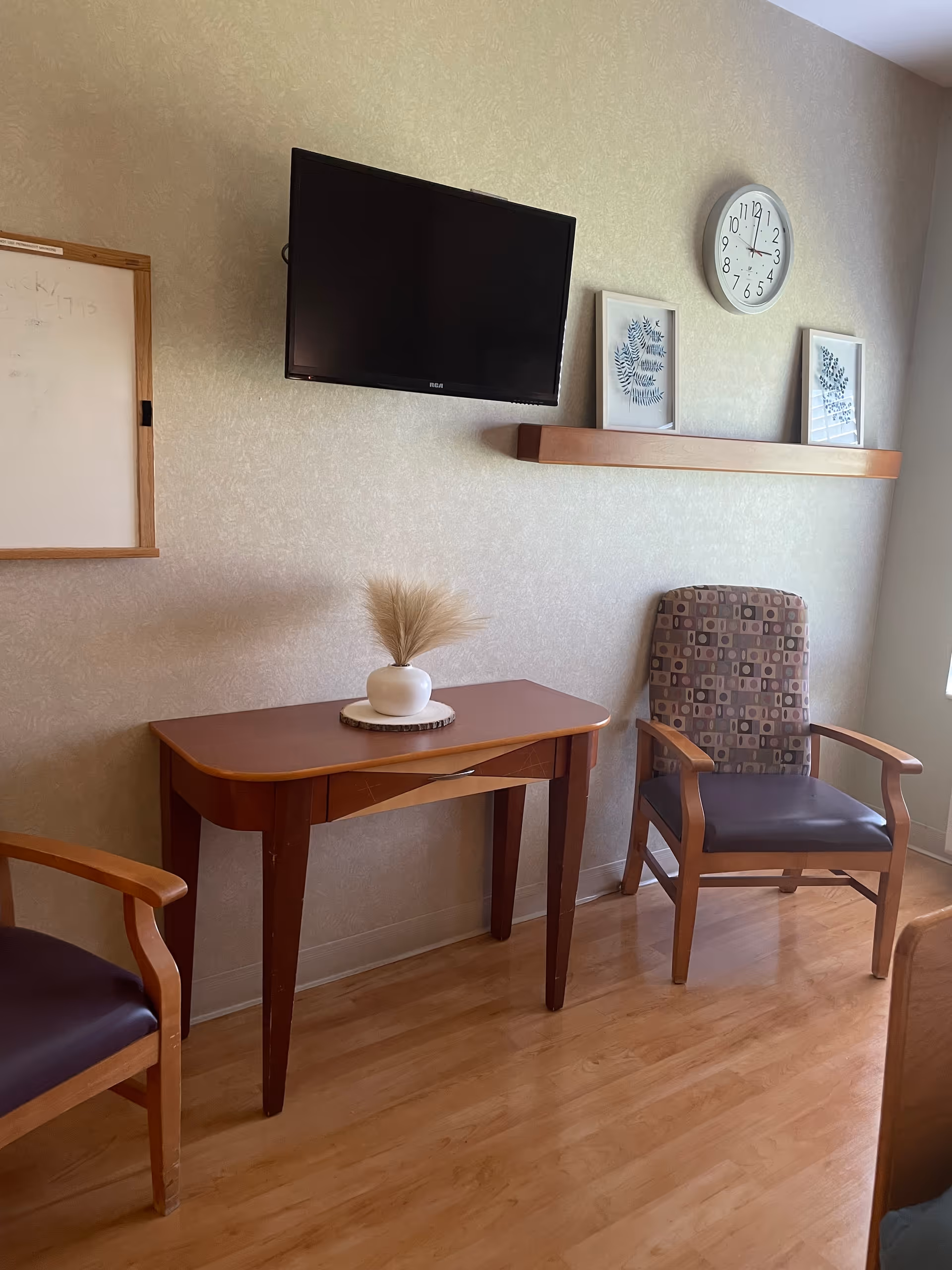 A small sitting area in a room with two wooden chairs with cushioned seats and backs, a wooden table with a small white vase holding dried grass, a wall-mounted flat screen TV, a whiteboard, a wall clock, and two framed botanical prints on a wooden shelf.